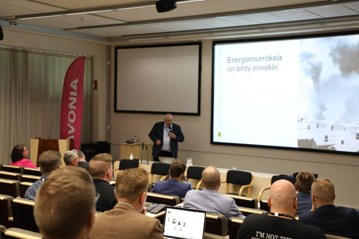 Pukuun pukeutunut mies pitää esityksen luokkahuoneessa; useita ihmisiä istuu häntä vastapäätä. Näytöllä olevassa diassa lukee Energiamurroksia on tehty ennenkin. Vasemmalla näkyy vaaleanpunainen Savonian banneri.