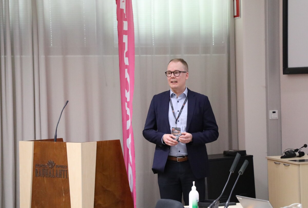 Pukuun pukeutunut mies seisoo lähellä puhujakoroketta juoma kädessään ja hymyillen kokoushuoneessa, jossa on mikrofoneja ja toimistolaitteita. Taustalla on vaaleanpunainen pystybanneri ja beigenväriset verhot.