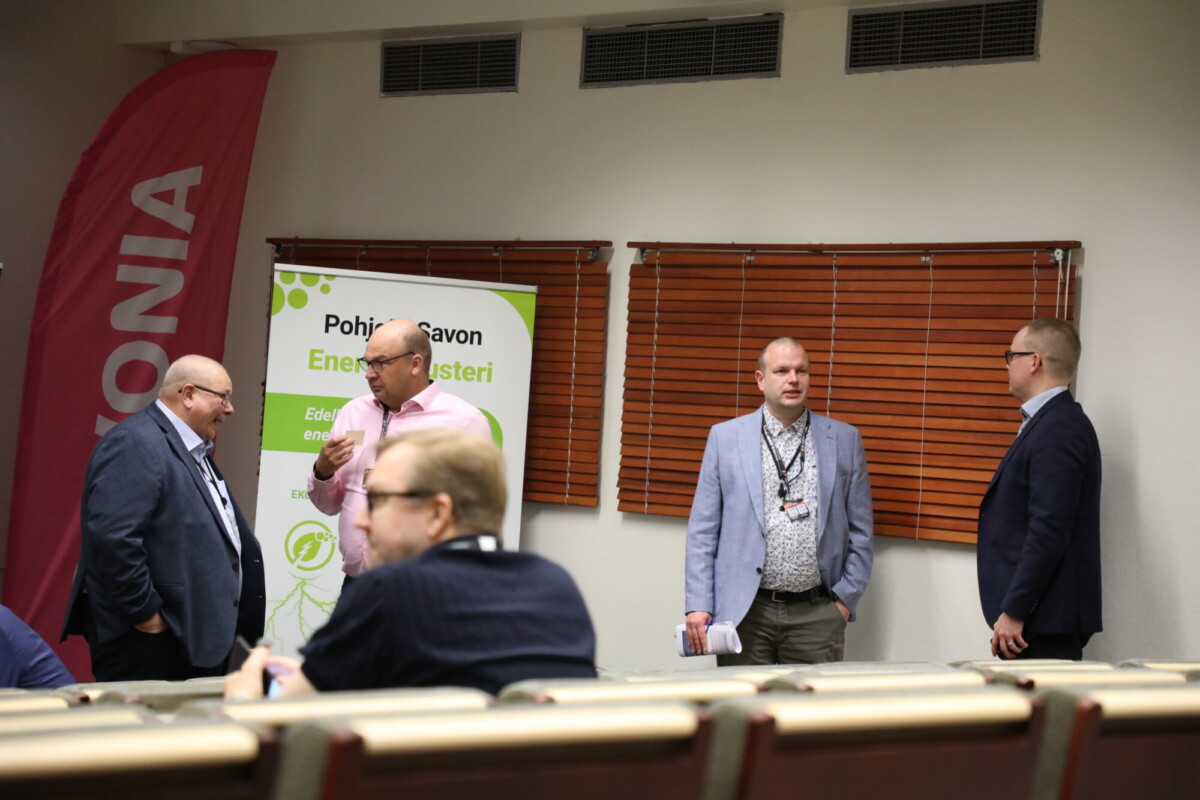 Neljä bisnesasuihin pukeutunutta miestä seisoo ja keskustelee kokoushuoneessa lähellä Pohjois-Savon Energian Klusteri -banneria. Etualalla on rivejä tyhjiä tuoleja; puiset kaihtimet peittävät ikkunoita.