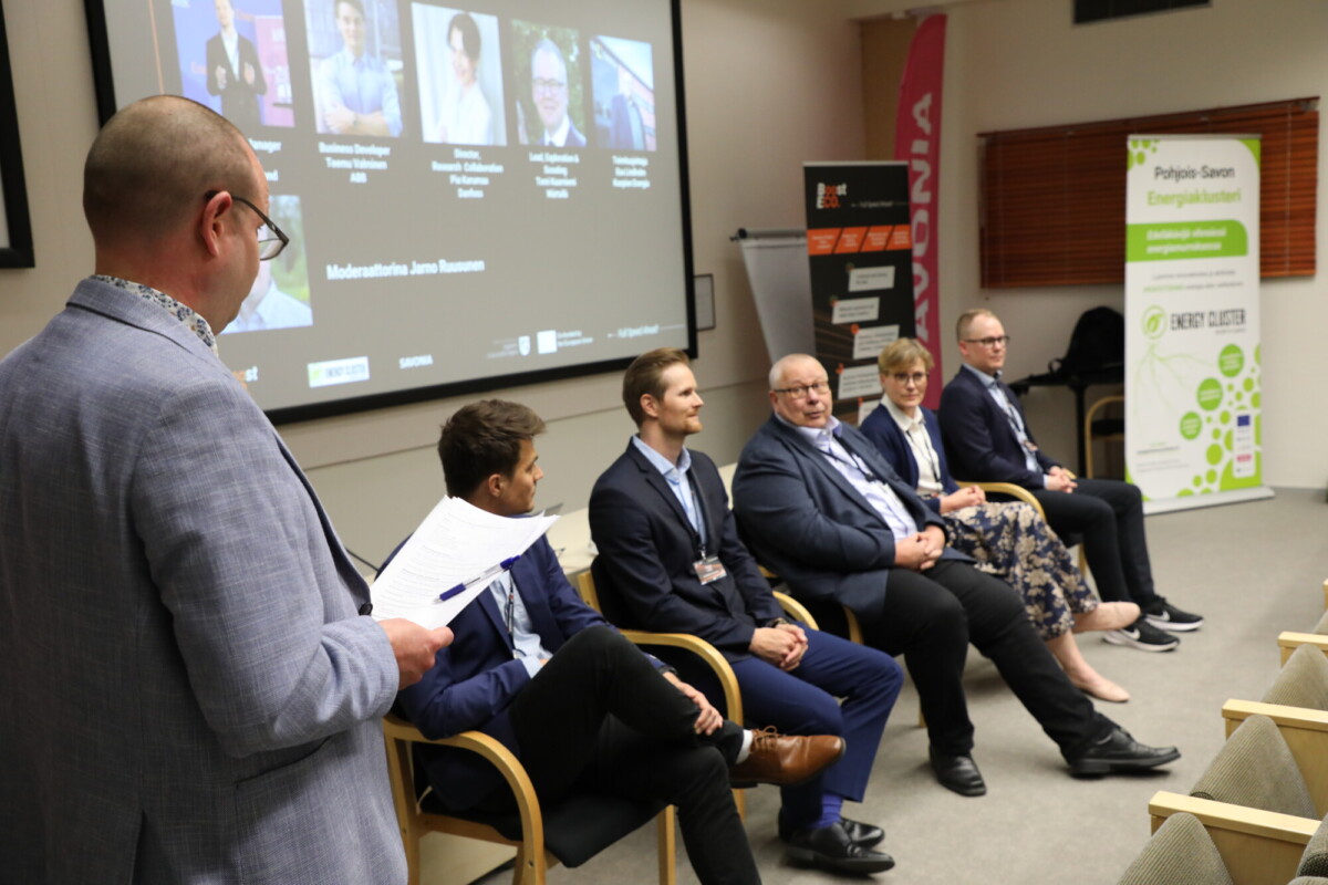 Mies seisoo papereita pitelemässä ja katsoo viittä istuvaa, bisnesasuihin pukeutunutta panelistia lavalla, jonka takana on suuri esitysnäyttö ja sivuilla tapahtumabannerit kokoushuoneessa.