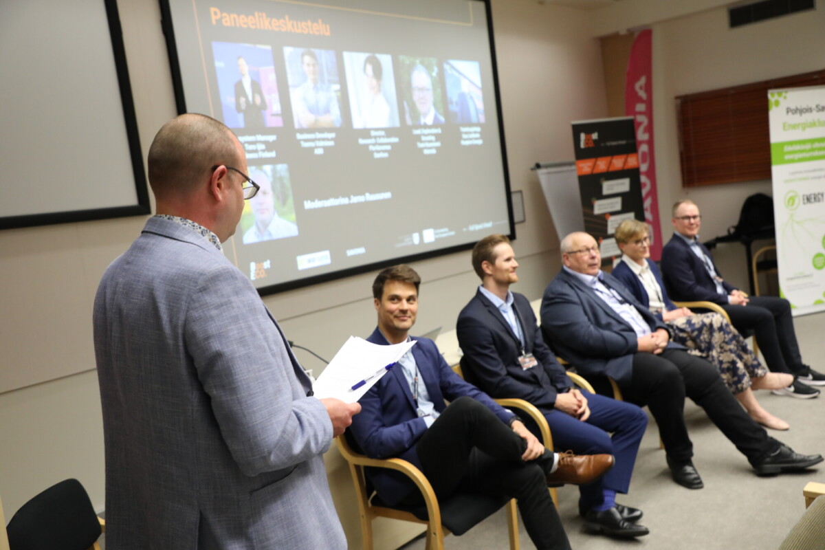 Moderaattori seisoo papereiden kanssa ja puhuu viidelle istuvalle, bisnesasuihin pukeutuneelle panelistille keskustelutilaisuuden aikana. Panelisteinien tiedot sisältävä esitysdia heijastetaan heidän taakseen olevalle näytölle.