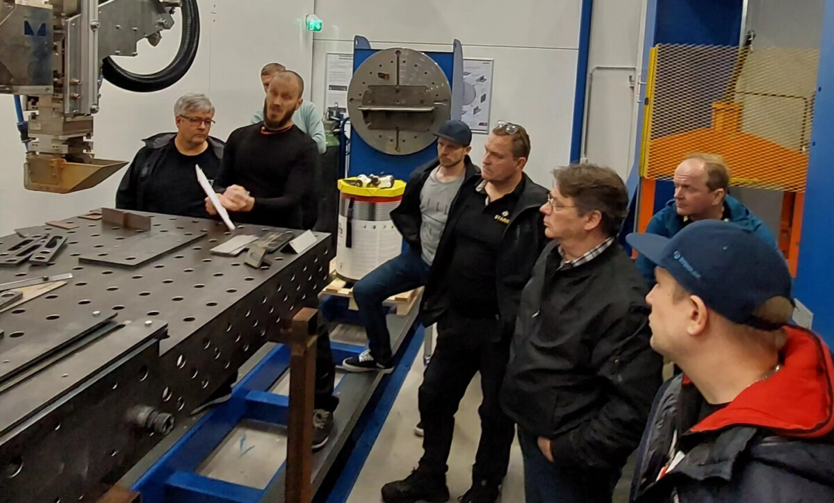 Ryhmä miehiä seisoo suuren metallisen työpöydän ympärillä teollisuustyöpajassa ja kuuntelee tarkkaavaisesti, kun yksi henkilö selittää jotakin valkoisella kepillä tai osoittimella. Taustalla näkyy teollisuuslaitteita ja -työkaluja.
