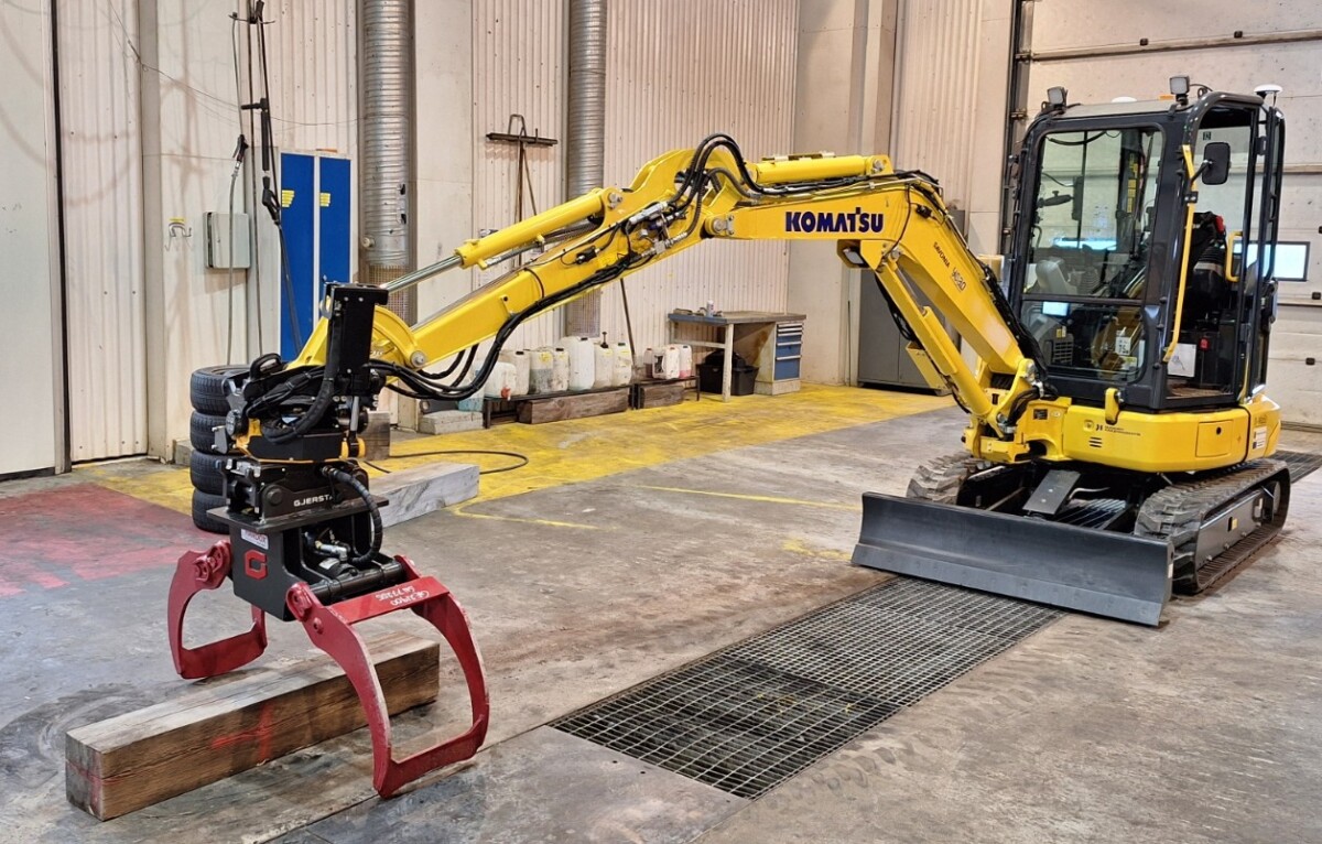 Keltainen kourakiinnitteinen Komatsu-minikaivuri nostaa puupalkkia tilavassa teollisuustyöpajassa, jossa on betonilattiat ja suuret ovet.