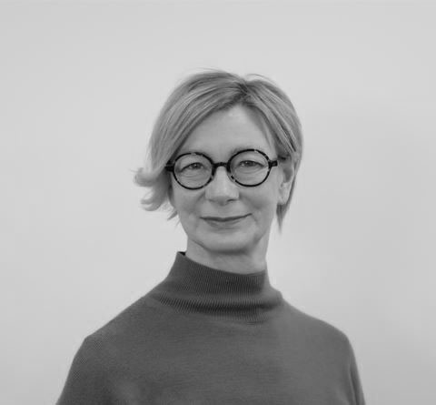 Lyhythiuksinen, vaaleatukkainen nainen, jolla on pyöreät mustat silmälasit ja korkeakauluksinen neule, poseeraa pehmeästi hymyillen yksinkertaista vaaleaa taustaa vasten mustavalkoisessa valokuvassa.