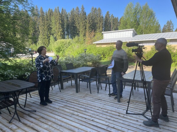 Nainen puhuu ja elehtii käsillään puisella terassilla, kun mies papereiden kanssa kuuntelee ja toinen mies kuvaa videokameralla. Taustalla näkyy vihreitä puita ja rakennuksia aurinkoisena päivänä.