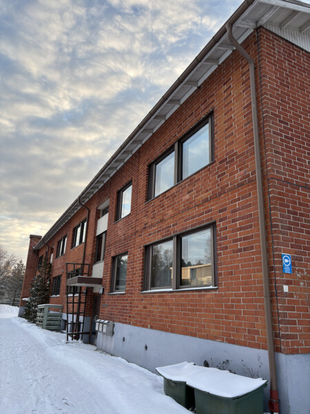 Kaksikerroksinen punatiilinen rakennus, jossa on suuret ikkunat ja edessä lumen peittämä jalkakäytävä, kuvattuna pilvisen taivaan alla talvella.