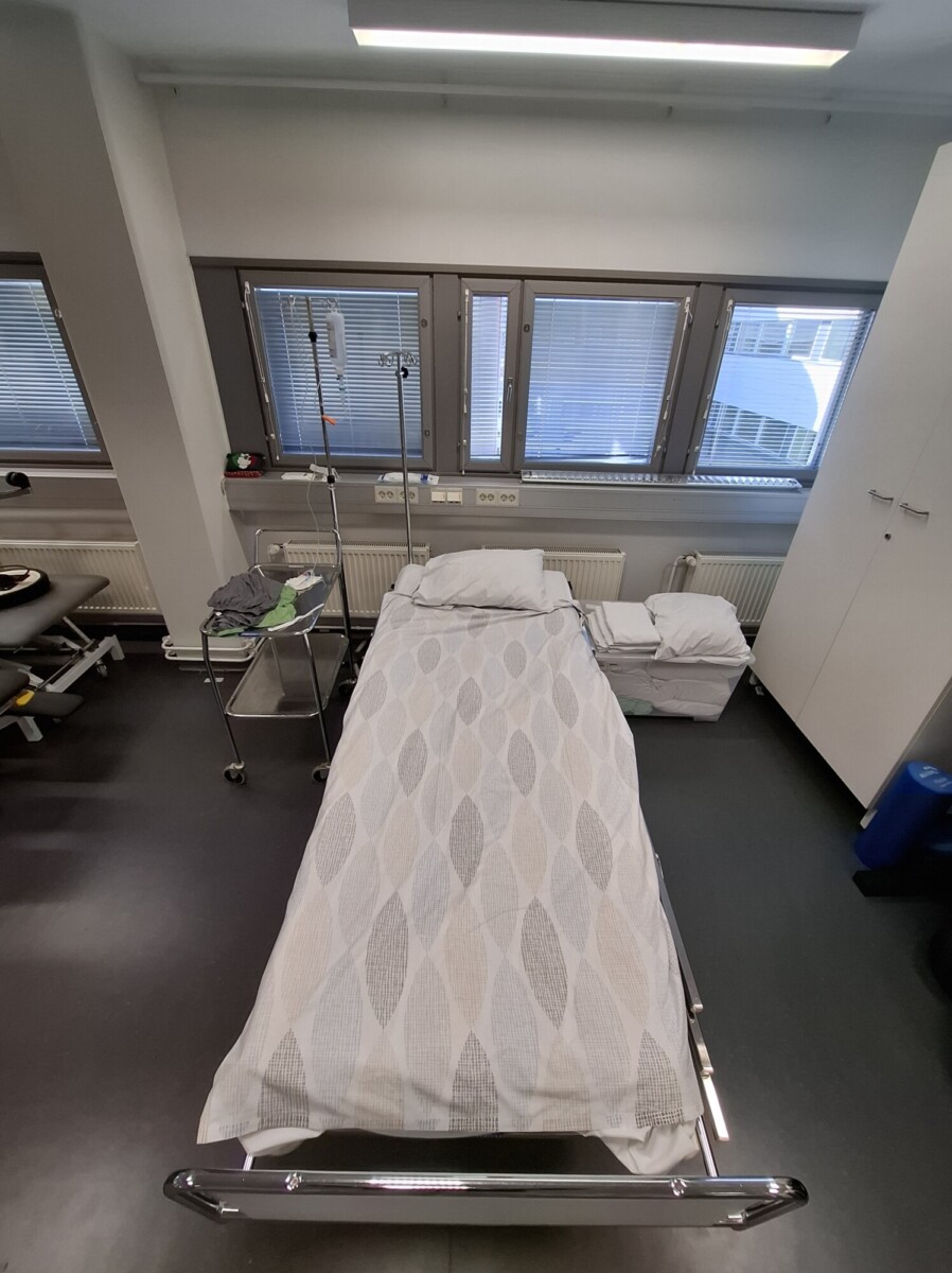Kuvioidun peiton peittämä sairaalasänky on sijoitettu ikkunan lähelle. Sänkyä ympäröivät lääketieteelliset laitteet, sivupöytä ja säilytyskaapit. Huone näyttää siistiltä ja järjestelmälliseltä.