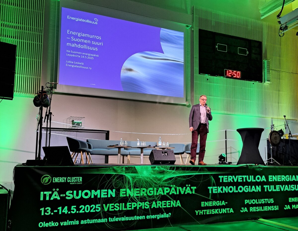 Mies seisoo lavalla puhumassa mikrofoniin energiakonferenssissa. Suurella näytöllä näkyy esitys nimeltä Energiamurros – Suomen suuri mahdollisuus. Tapahtuman tiedot näkyvät lavabannerissa.