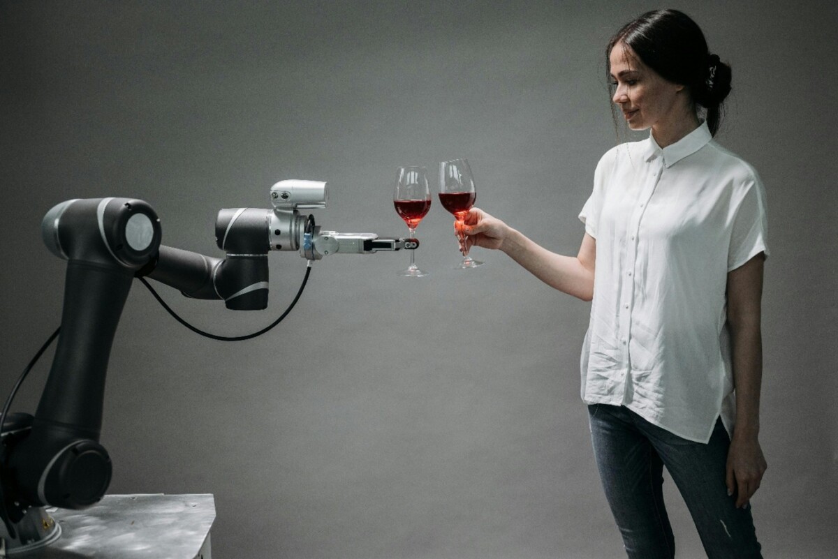 Nainen hymyilee ja kilisee viinilasia robottikäsivarrellaan, ja molemmat pitelevät lasillista punaviiniä yksinkertaista harmaata taustaa vasten.