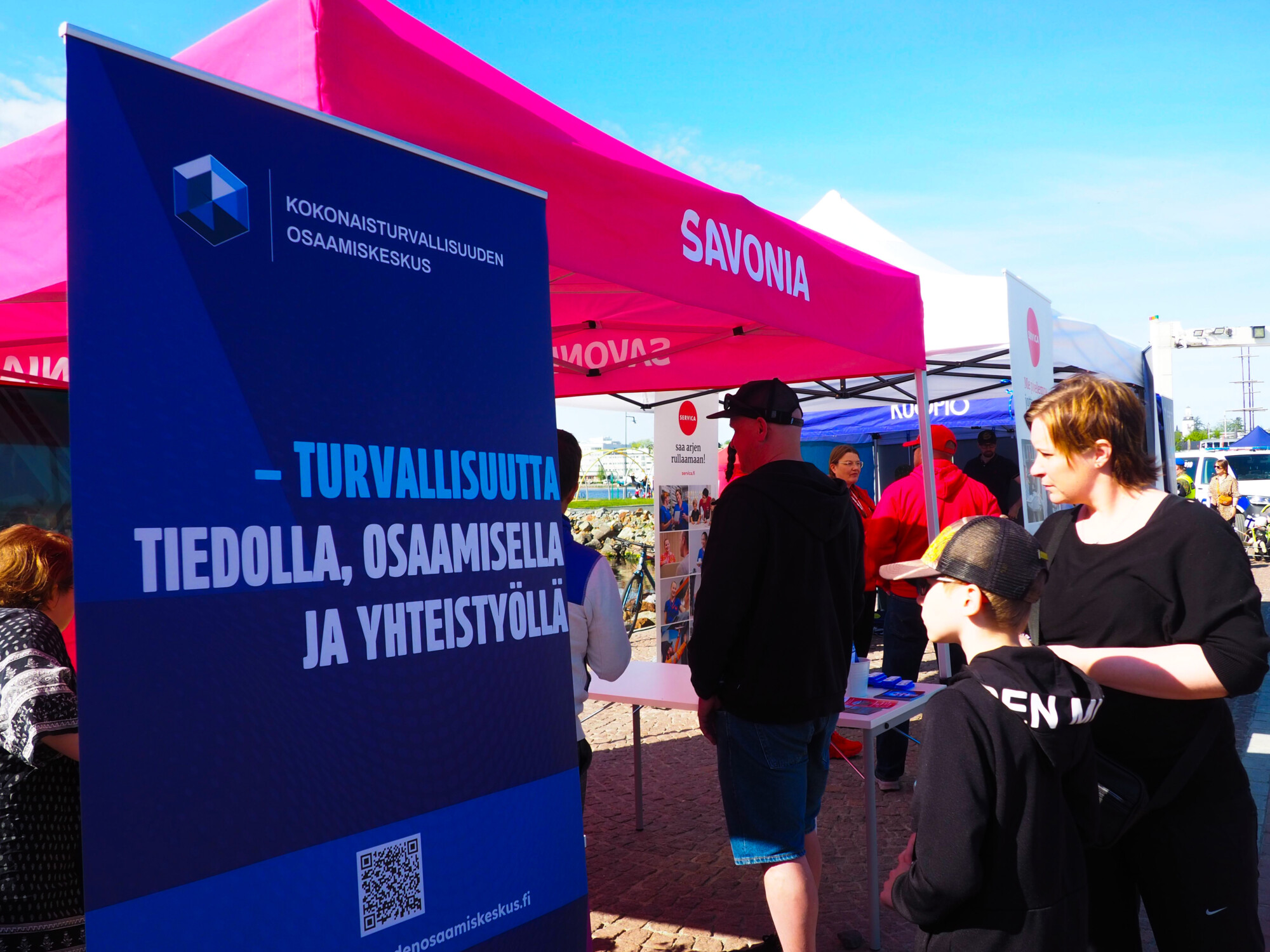 Ihmiset seisovat vaaleanpunaisten ja sinisten SAVONIA-tekstillä varustettujen infopisteiden lähellä. Suuri suomenkielinen kyltti edistää turvallisuutta tiedon, taitojen ja yhteistyön avulla. Tapahtuma järjestetään ulkona aurinkoisena päivänä.