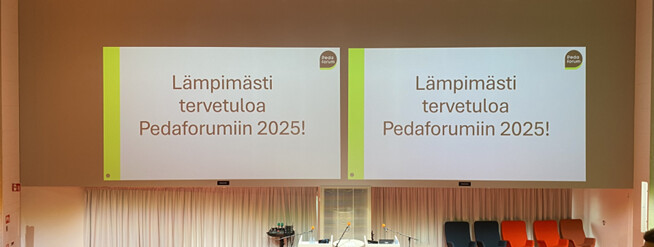 Auditorion suurilla valkokankailla näkyy viesti Lämpömästi tervetuloa Pedaforumiin 2025!, ja valkokankaiden alla näkyvät tuolit ja pöytä.
