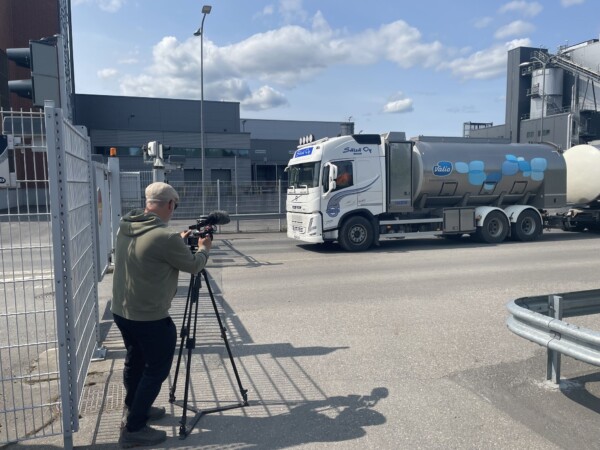 Henkilö käyttää jalustalla olevaa videokameraa tien varrella ja kuvaa Valio-merkkistä säiliöautoa, joka ajaa teollisuusrakennuksen ohi osittain pilvisellä taivaalla.