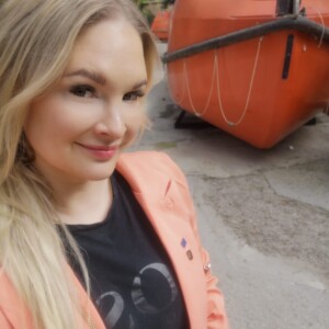 Pitkät vaaleatukkainen nainen, jolla on yllään persikanvärinen bleiseri ja musta t-paita, hymyilee ulkona selfietä varten. Hänen takanaan on kaksi suurta, oranssia, suljettua pelastusvenettä päällystetyllä alueella. Taustalla näkyy puita ja vehreyttä.