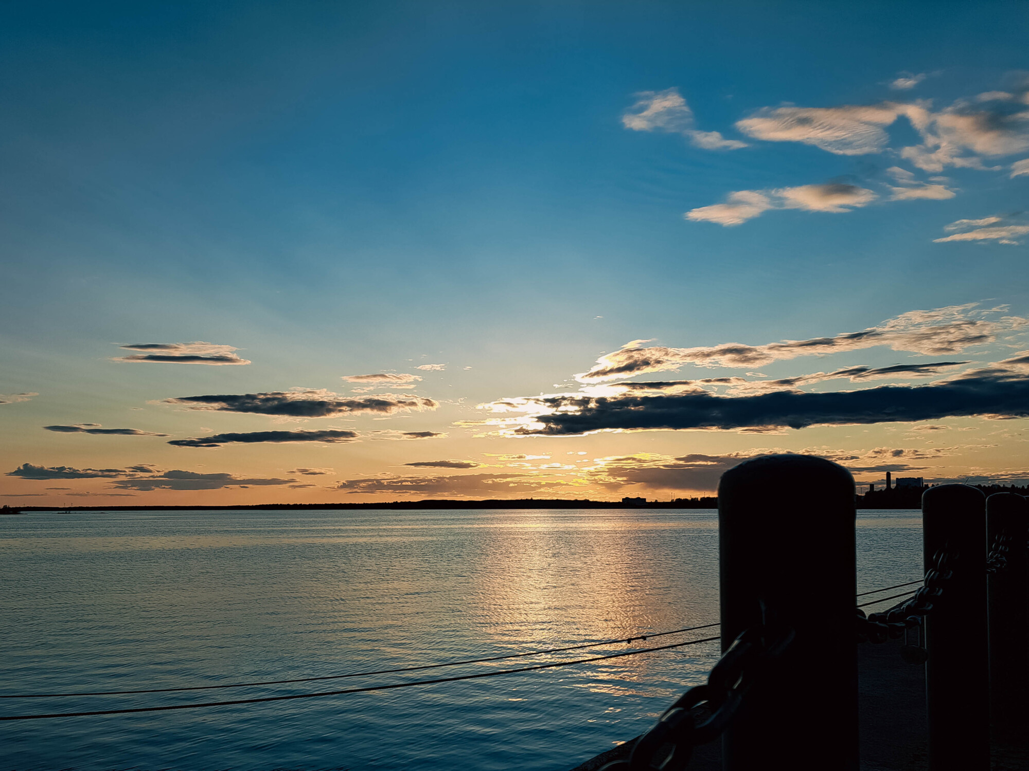 Auringonlasku tyynen vesistön yllä, pilviä hajallaan taivaalla. Etualalla rantaviivaa reunustavat mustat pylväät ja ketjut.