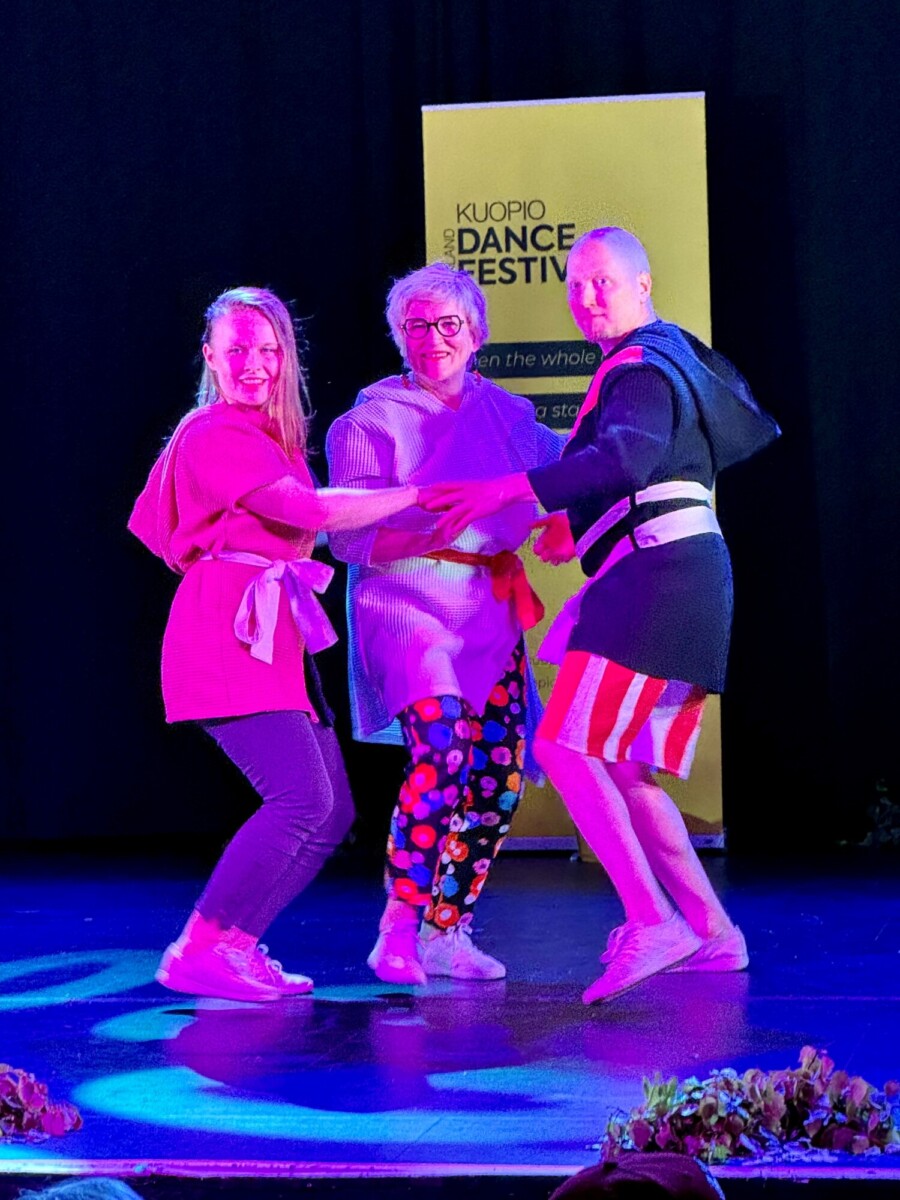 Kolme ihmistä hymyilee ja tanssii lavalla violetin valaistuksen alla. He pitävät kädestä kiinni ympyrässä. Taustalla on kirkkaan keltainen "Kuopio Dance Festival" -banderoli. Tanssijoilla on yllään värikkäät, rennot vaatteet.
