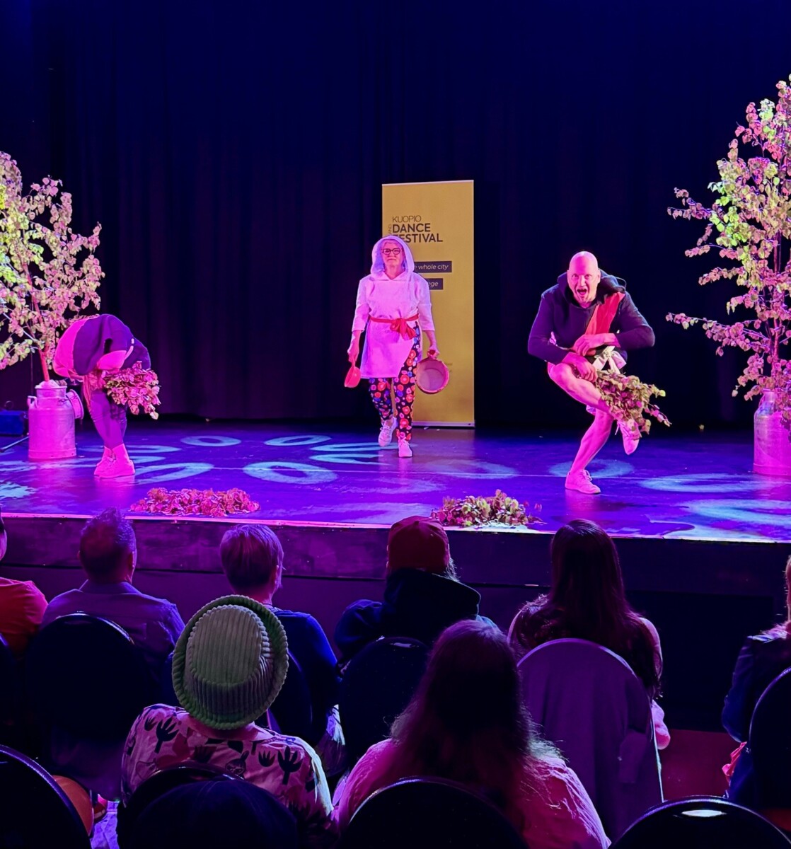 Kolme esiintyjää lavalla yleisön edessä tanssimassa oksien kanssa violetin valaistuksen alla ja kahden kukka-asetelman vieressä. Heidän takanaan on keltainen kyltti, jossa lukee Kansantanssifestivaali.