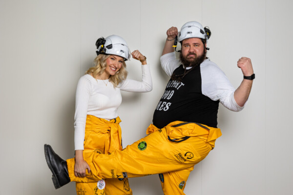 Kaksi keltaisiin haalareihin ja valkoisiin kypäriin pukeutunutta ihmistä poseeraa leikkisästi heilutellen käsiään ja hymyillen kameralle. Toisella on valkoinen paita, toisella musta paita, jossa on valkoista tekstiä.