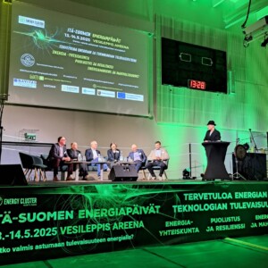 Kuusi puvuissa olevaa miestä istuu lavalla korokkeen vieressä, jossa mustapukuinen mies puhuu. Vihreissä valoissa ja banderolleissa lukee ”Itä-Suomen Energiapäivät” ja tapahtuman tietoja futuristista graafista taustaa vasten.