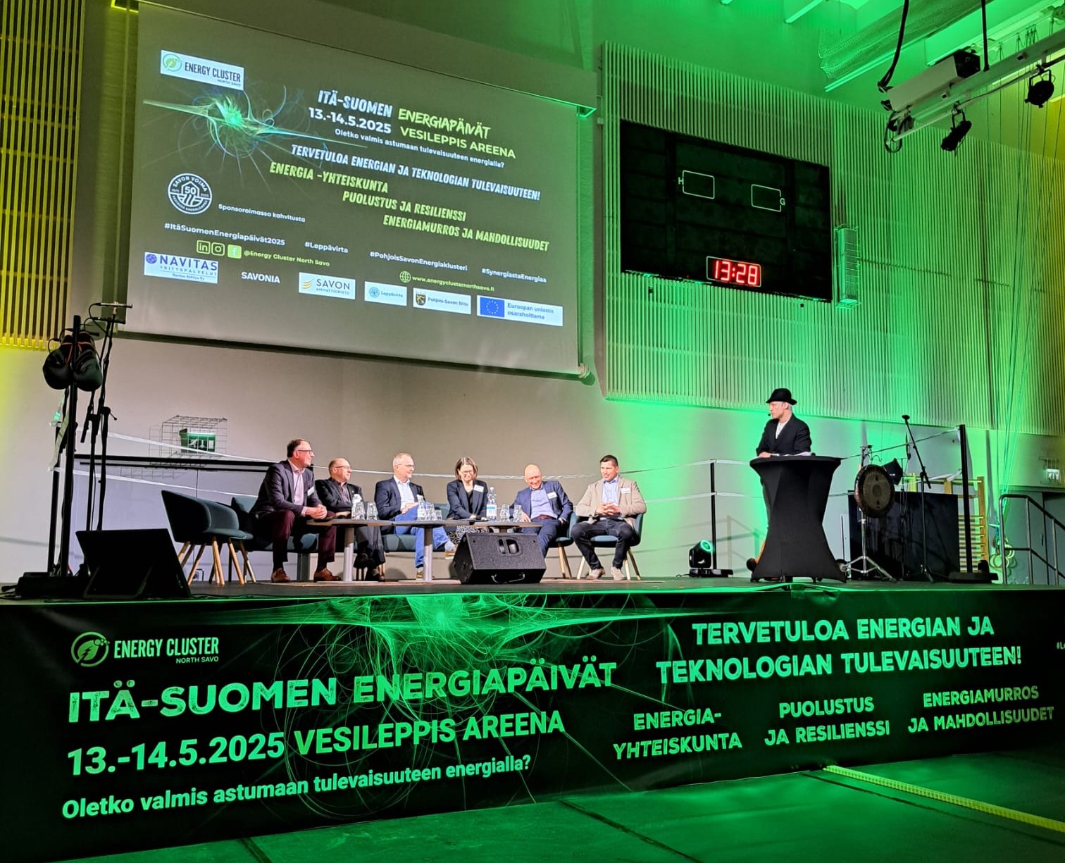 Kuusi puvuissa olevaa miestä istuu lavalla korokkeen vieressä, jossa mustapukuinen mies puhuu. Vihreissä valoissa ja banderolleissa lukee ”Itä-Suomen Energiapäivät” ja tapahtuman tietoja futuristista graafista taustaa vasten.