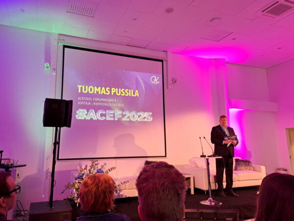 Pukuun pukeutunut mies seisoo lavalla valkoisen sohvan vieressä pitämässä esitystä. Hänen takanaan olevalla näytöllä lukee teksti Tuomas Pussila ja #ACEF2025 huoneessa, joka on valaistu violeteilla ja vaaleanpunaisilla valoilla.