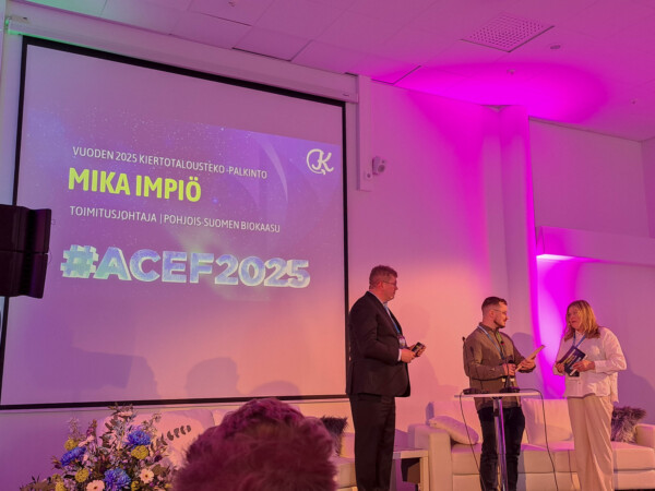 Kolme ihmistä seisoo lavalla palkintojenjakotilaisuudessa. Heidän takanaan olevalla näytöllä lukee ”Vuoden 2025 Kiertotalousteko - Palkinto Mika Impiö” ja ”#ACEF2025”. Lava on valaistu violetilla valolla ja koristeltu kukilla.