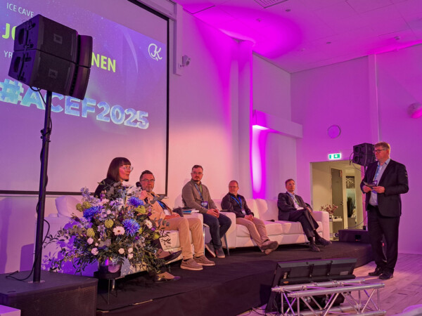 Kuuden hengen paneeli istuu lavalla konferenssissa, ja yksi henkilö seisoo puhumassa yleisölle. Taustalla on teksti "#ACEF2025" ja huone on valaistu violetilla valolla. Edessä näkyy kukka-asetelma.