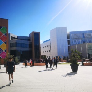Opiskelijat kävelevät ja kokoontuvat aurinkoisella sisäpihalla modernien yliopistorakennusten välissä. Yhdessä rakennuksessa on värikäs geometrinen seinämaalaus, kun taas toisissa on suuret ikkunat ja valkoiset julkisivut. Ruukkukasvit koristavat aluetta.