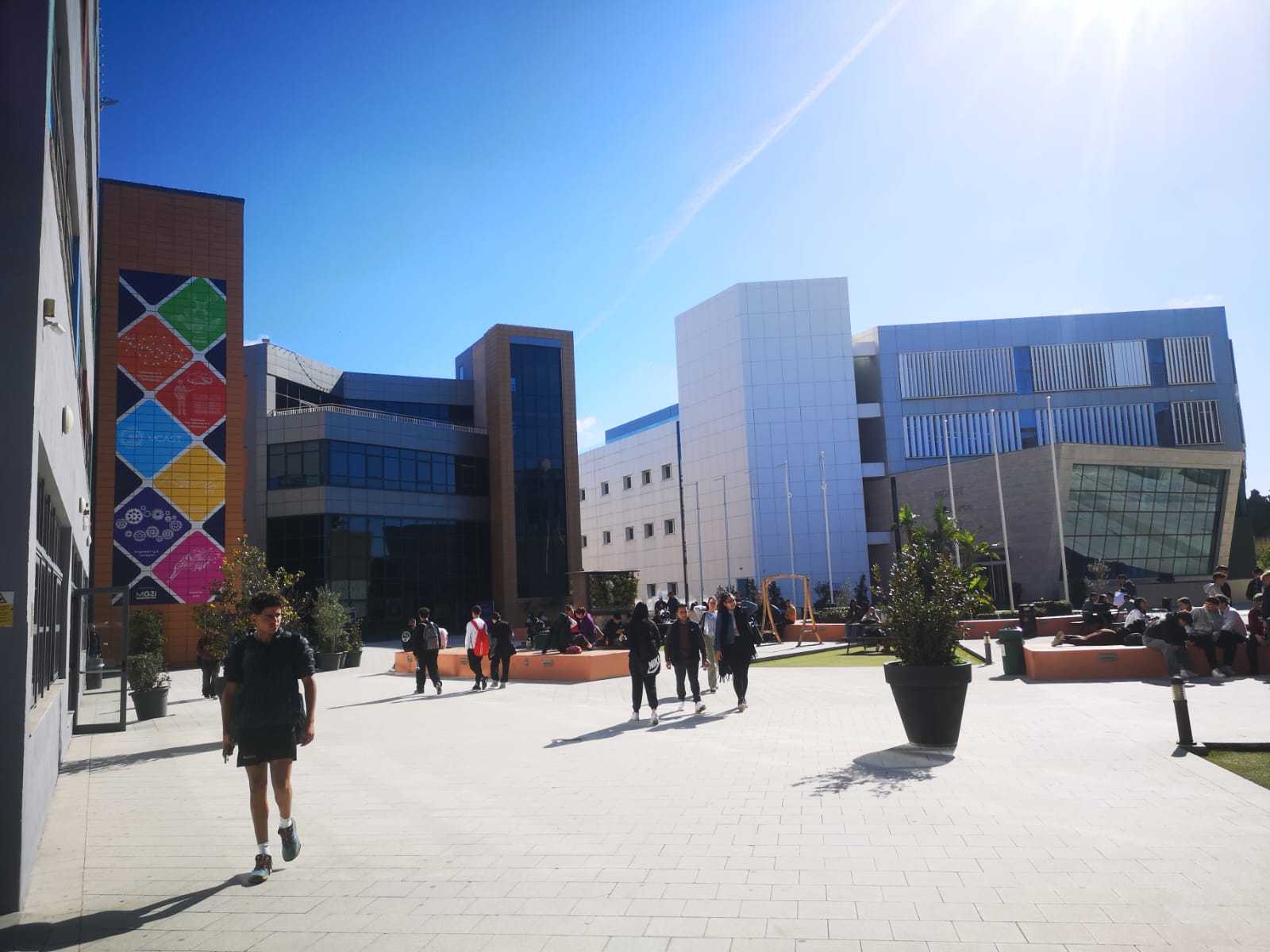 Opiskelijat kävelevät ja kokoontuvat aurinkoisella sisäpihalla modernien yliopistorakennusten välissä. Yhdessä rakennuksessa on värikäs geometrinen seinämaalaus, kun taas toisissa on suuret ikkunat ja valkoiset julkisivut. Ruukkukasvit koristavat aluetta.