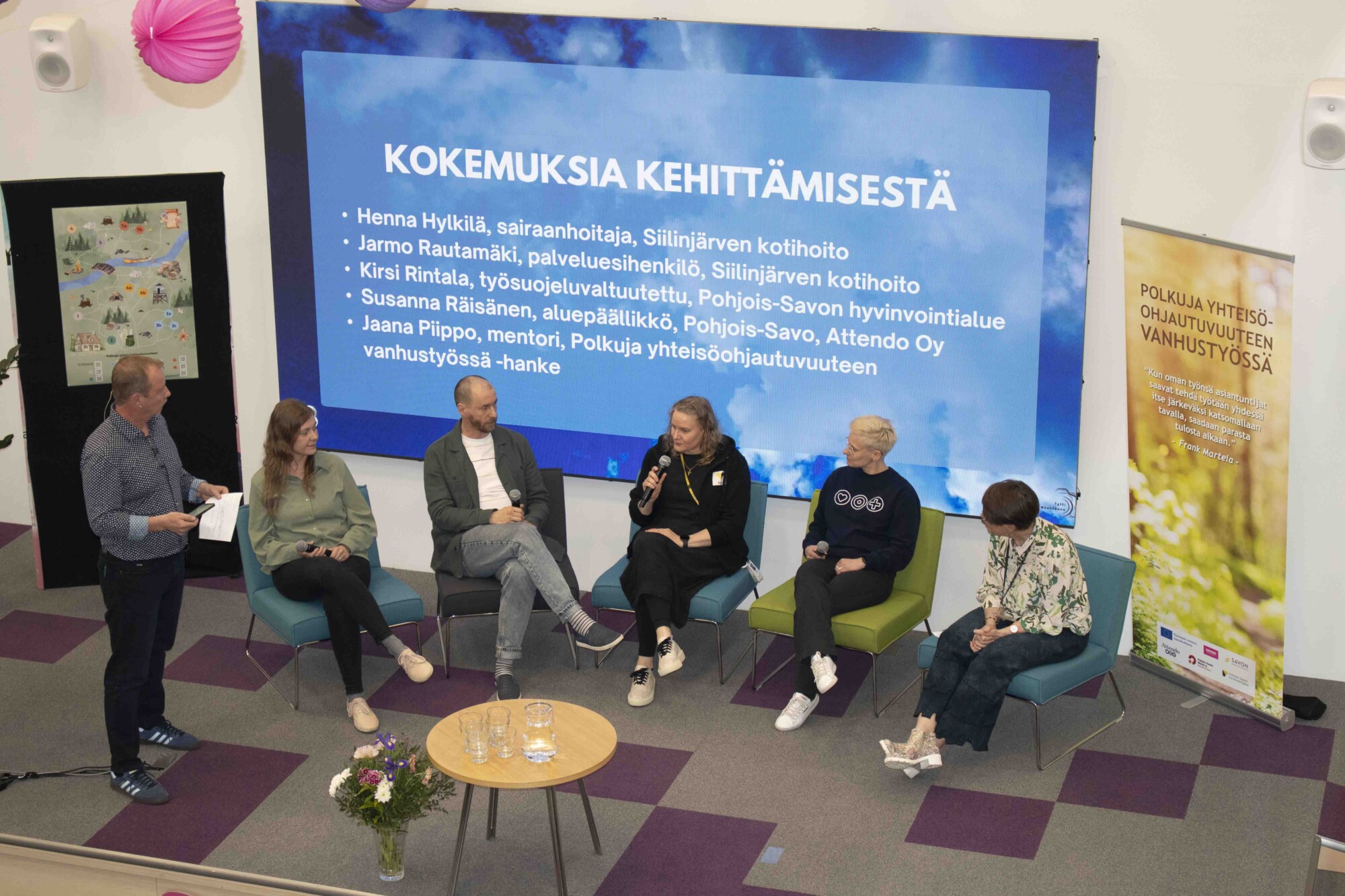 Viisi ihmistä istuu lavalla nojatuolissa paneelikeskustelun aikana. Vasemmalla seisoo mies mikrofonin kanssa. Heidän takanaan on suuri näyttö, jossa on suomenkielistä tekstiä ja banneri. Panelistien edessä pienellä pöydällä on kukkia.