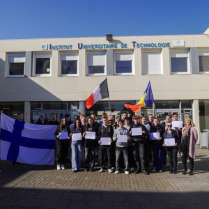 Ryhmä ihmisiä seisoo Institut Universitaire de Technologie -rakennuksen ulkopuolella kirkkaan sinisen taivaan alla. Joillakin on hallussaan Suomen, Ranskan ja Romanian todistuksia ja lippuja.