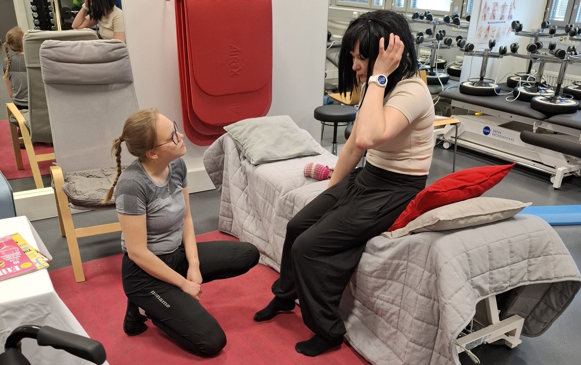 Nainen istuu sängyllä kuntosalilla tai klinikalla ja koskettaa päätään, kun taas toinen nainen polvistuu hänen viereensä ja tarjoaa tukea. Taustalla näkyvät liikuntavälineet ja kaaviot.