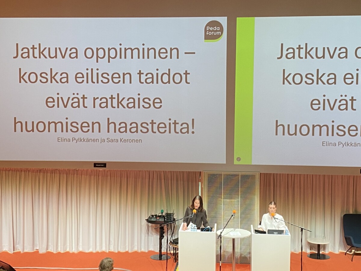 Kaksi naista seisoo erillisillä korokkeilla lavalla ja esittelee suuren valkokankaan edessä, jolla on suomenkielinen teksti jatkuvasta oppimisesta. Tapahtuma vaikuttaa muodolliselta, ja yleisöä on todennäköisesti paikalla.
