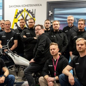 Viidentoista hengen ryhmä poseeraa sisätiloissa moottoripyörän ympärillä. Useimmilla on yllään mustat paidat tai hupparit, joissa on teksti SAVONIA. Yksi henkilö pitelee mustaa t-paitaa, jossa on valkoiset vaihdekuvakkeet. Kyltissä lukee SAVONIA KONETEKNIIKKA.