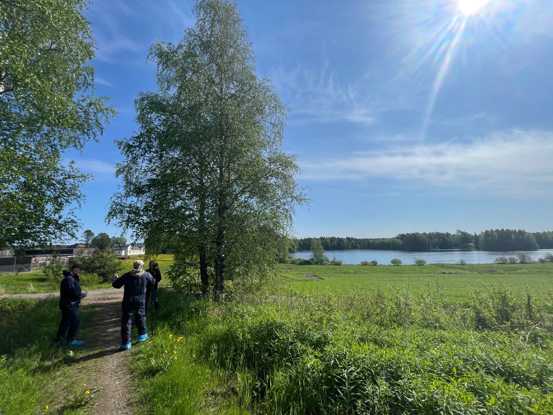 Kolme ihmistä seisoo ruohikkoisella polulla korkeiden puiden vieressä kirkkaassa auringossa ja sinisellä taivaalla, josta avautuu näkymä rauhalliselle joelle ja vihreille pelloille kaukana.