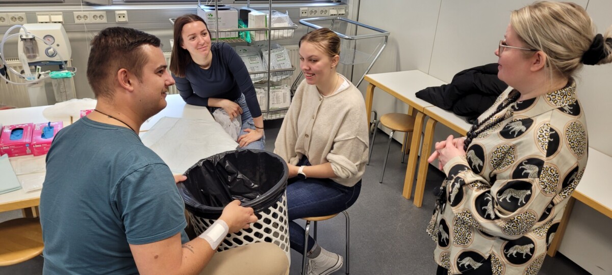 Neljä ihmistä on kokoontunut luokkahuoneeseen tai laboratorioon; yksi mies pitelee avointa roska-astiaa, kun taas kolme naista, kaksi istuu ja yksi seisoo, katsovat ja ovat vuorovaikutuksessa hänen kanssaan. Taustalla näkyy lääkintätarvikkeita.