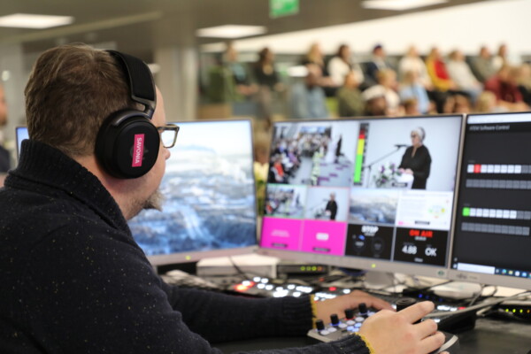 Henkilö, jolla on kuulokkeet, käyttää videotuotantolaitteita ohjauspöydän ääressä ja valvoo useita näyttöjä, joilla näytetään suoraa tapahtumaa, jonka taustalla on puhuja ja yleisö.