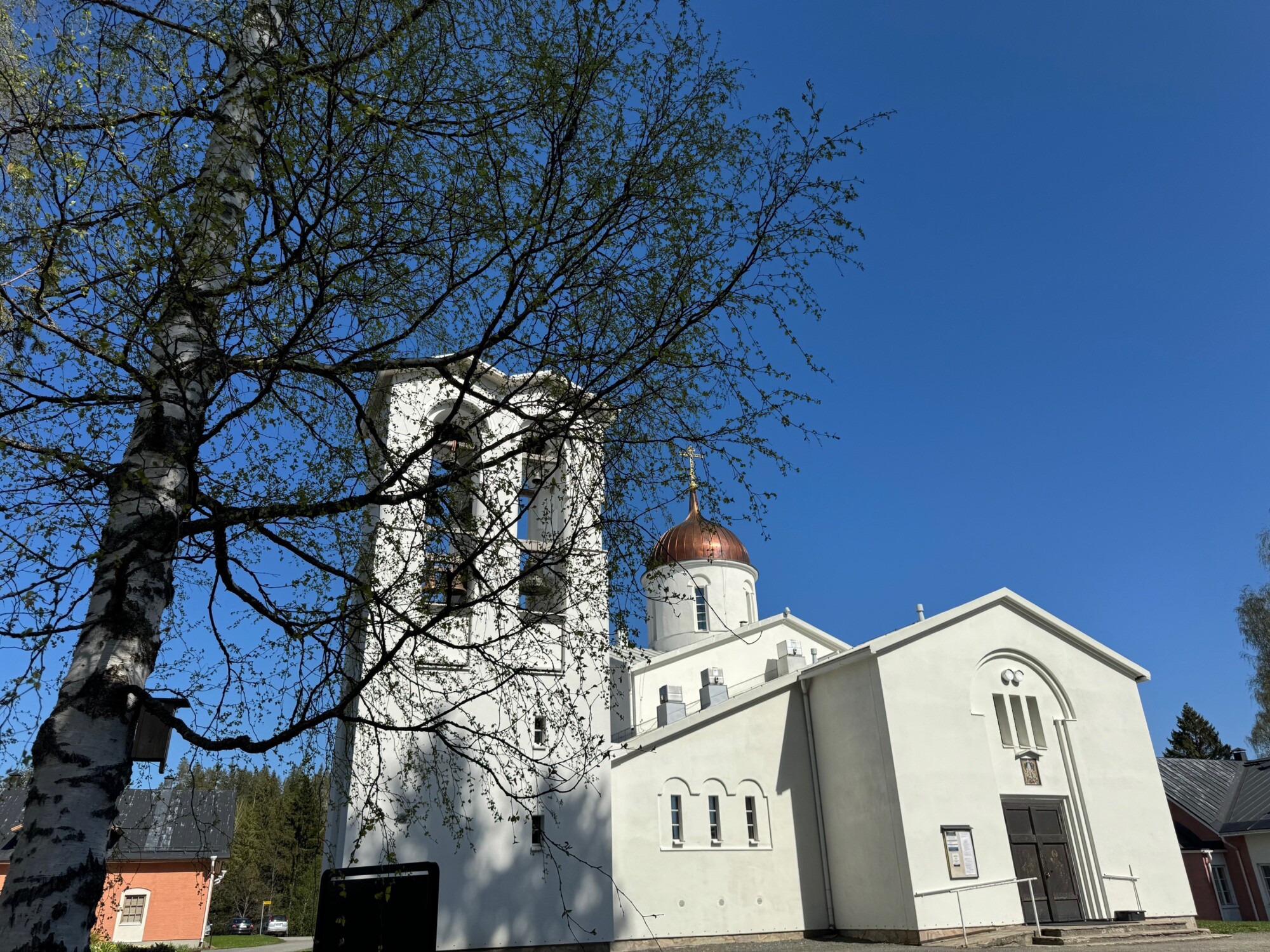 Valkoinen kirkko, jossa on kellotorni ja kuparikupoli, seisoo kirkkaansinisen taivaan alla. Etualalla on nuorta lehtipuuta kasvava koivu, ja taustalla näkyy muita rakennuksia ja puita.
