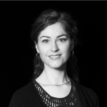 Mustavalkoinen muotokuva naisesta, jolla on aaltoilevat hiukset, korvakorut ja tumma toppi, jossa on kuvioitu kaula-aukko, ja joka hymyilee pehmeästi tummaa taustaa vasten.