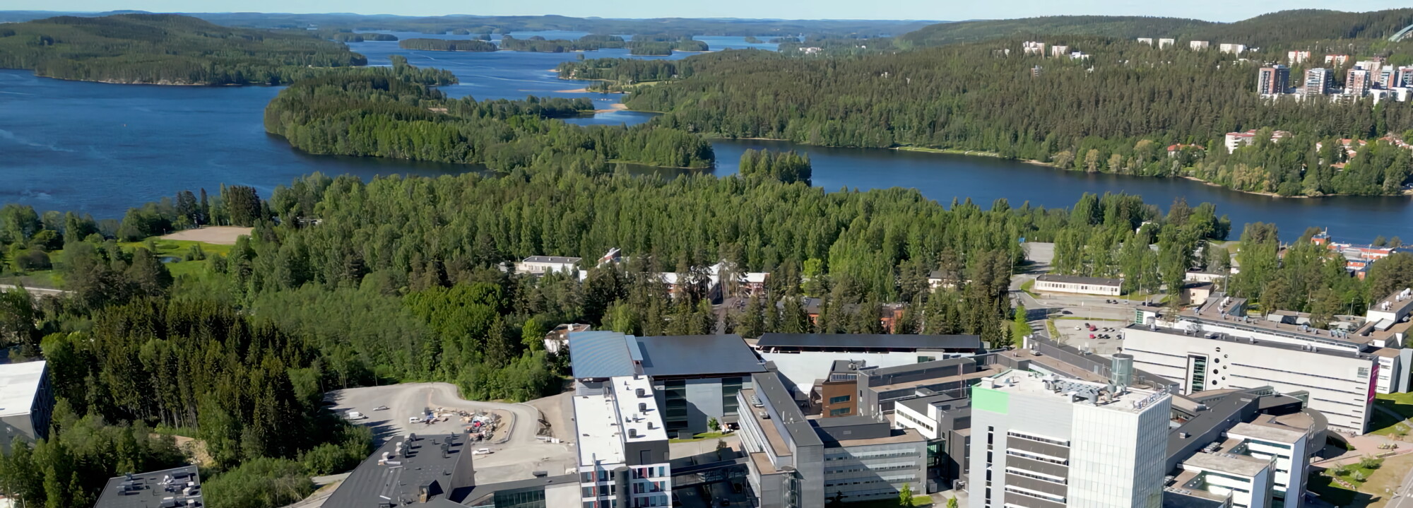 Ilmakuva modernista kampuksesta, jota ympäröi tiheä metsä ja järvet, etualalla useita monikerroksisia rakennuksia ja taustalla maisemallinen vesi ja vihreät kukkulat.