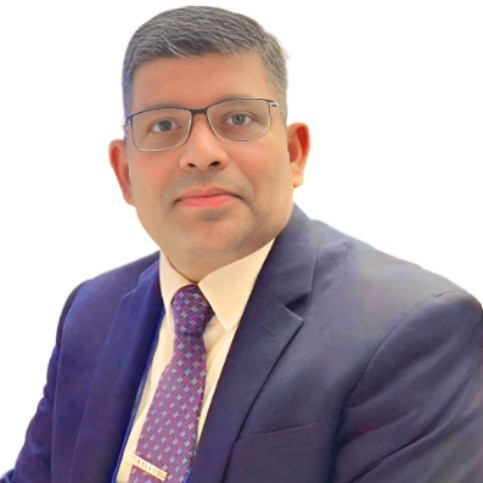 Mies, jolla on silmälasit, tumma puku, valkoinen paita ja kuviollinen solmio, istuu kameraan päin valkoista taustaa vasten.