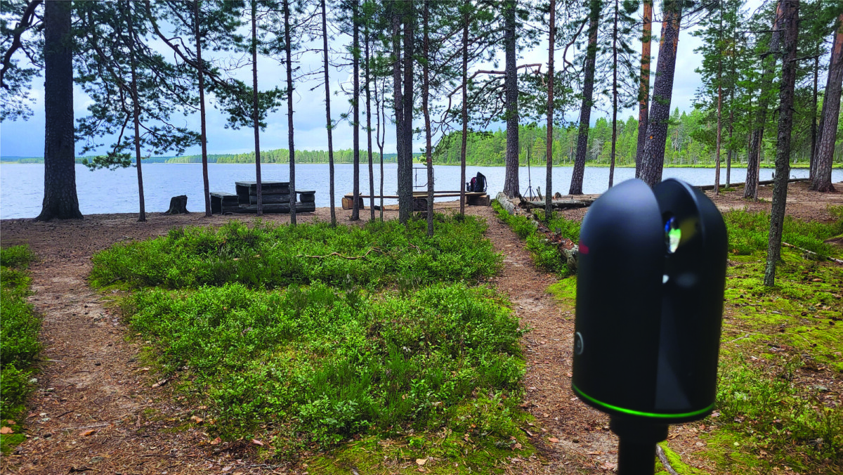 Jalustalla oleva musta kamera seisoo metsäaukiolla järven lähellä. Paikalla on piknikpöytiä, penkkejä ja korkeita mäntyjä, ja taustalla näkyy vettä ja kaukaisia puita.
