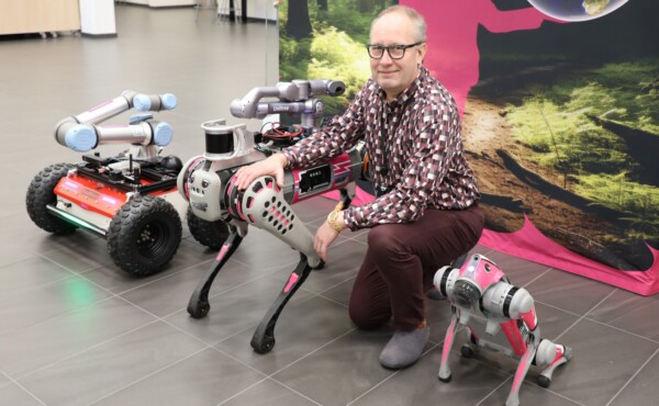 Mies polvistuu sisätiloissa kolmen robotin vieressä: robottikoira, pyörillä liikkuva robottikäsi ja pieni robottikoira. Taustalla on luontoaiheinen seinänäyttely.