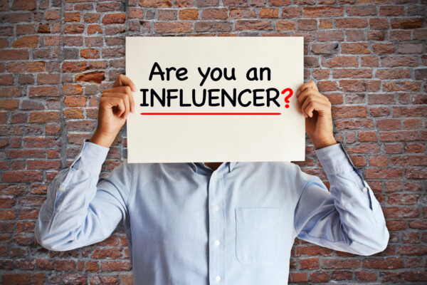 Vaaleansiniseen paitaan pukeutunut henkilö seisoo tiiliseinää vasten ja pitää kasvojensa edessä kylttiä, jossa lukee Are you an INFLUENCER? ja jossa on alleviivattuna INFLUENCER? ja punainen kysymysmerkki.