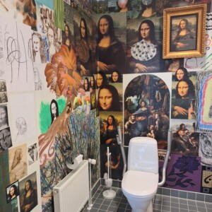 Kylpyhuone, jossa on harmaa laattalattiat ja seinät, jotka on peitetty erilaisilla Mona Lisa -taideteoksilla, luonnoksilla ja jäljennöksillä. Valkoinen wc ja WC-harja ovat näkyvissä, ja vasemmalla seinällä on patteri.