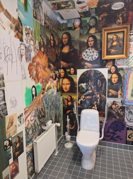 Kylpyhuone, jossa on harmaa laattalattiat ja seinät, jotka on peitetty erilaisilla Mona Lisa -taideteoksilla, luonnoksilla ja jäljennöksillä. Valkoinen wc ja WC-harja ovat näkyvissä, ja vasemmalla seinällä on patteri.