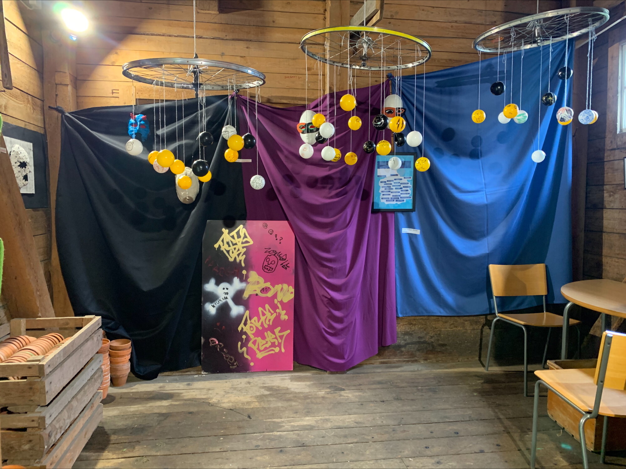 Viihtyisä puinen huone, jonka taustana on mustaa, violettia ja sinistä kangasta. Katosta roikkuvat polkupyöränpyörät, jotka on koristeltu keltaisilla, valkoisilla ja mustilla palloilla. Paikalla on pinottuja tuoleja, pöytä ja laatikoita kukkaruukkuineen.