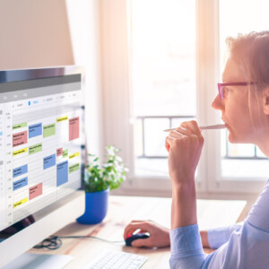 Silmälasipäinen nainen istuu työpöydän ääressä, katsoo mietteliäästi tietokoneen näyttöä, jossa näkyy värikäs digitaalinen kalenteri, ja pitää kynää suullaan. Ikkunan vieressä olevalla pöydällä on pieni kasvi ja lamppu.