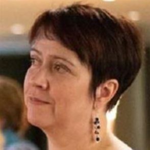 Nainen, jolla on lyhyet tummat hiukset ja roikkuvat korvakorut, katsoo hieman vasemmalle. Tausta on pehmeästi sumentunut lämpimällä valaistuksella.