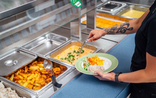 Henkilö tarjoilee ruokaa kahvilan noutopöydästä, lautasella on riisiä ja vihanneksia. Noutopöydän tarjottimilla on paahdettuja perunoita, yrttien kanssa kuorrutettua kermaista ruokaa ja muita lämpimiä ruokia.