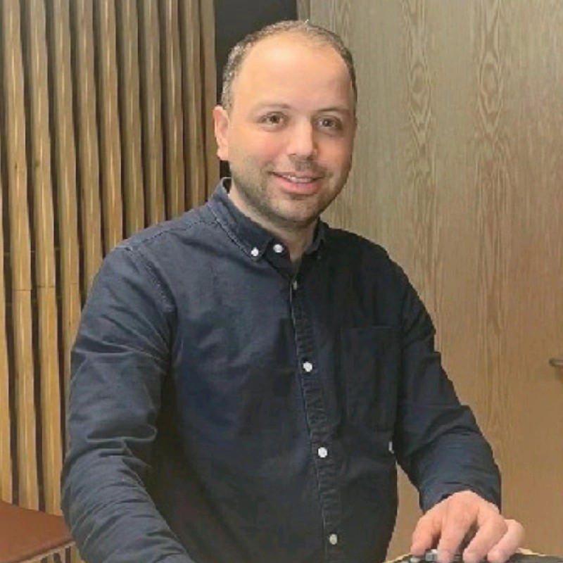 Lyhythiuksinen mies, jolla on tumma nappipaita, seisoo sisätiloissa pystysuorilla säleillä varustetun puuseinän edessä ja hymyilee kameralle.